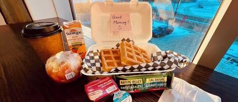 Bandeja de desayuno con waffles, jugo de naranja, una barra de pan y una caja de bocadillos Nature Valley dulces y salados sobre una mesa junto a una ventana con vistas nevadas.