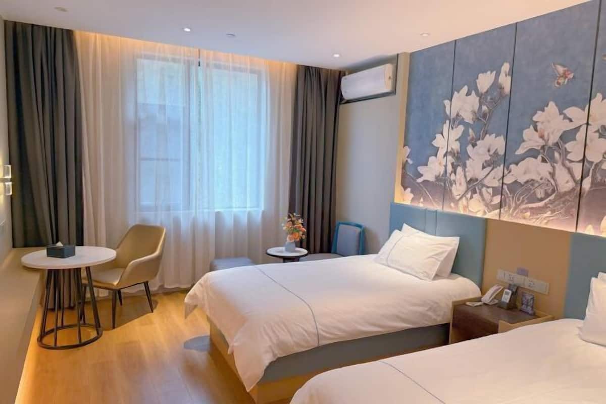 A hotel room with two beds, a small table, a chair, a window with curtains, and a wall with a floral painting.
