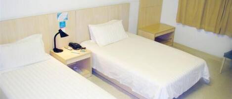 A hotel room with two single beds, a bedside table with a lamp, a small shelf, and a wall-mounted coat rack.
