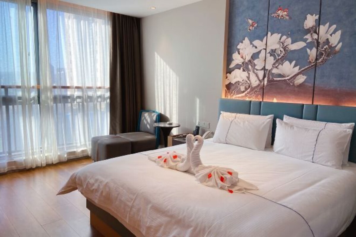 Una habitación de hotel con una cama grande, un cuadro de flores en la pared y un balcón con cortinas.