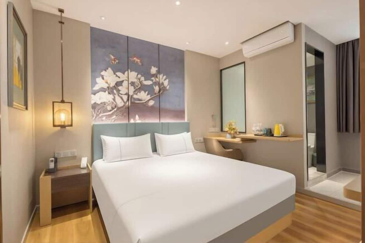 A modern hotel room with a large bed, a desk, a chair, a painting, and a mirror.