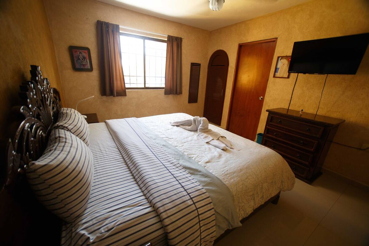 Habitación de hotel con una cama grande, un aparador de madera, televisor montado en la pared y una ventana con cortinas.