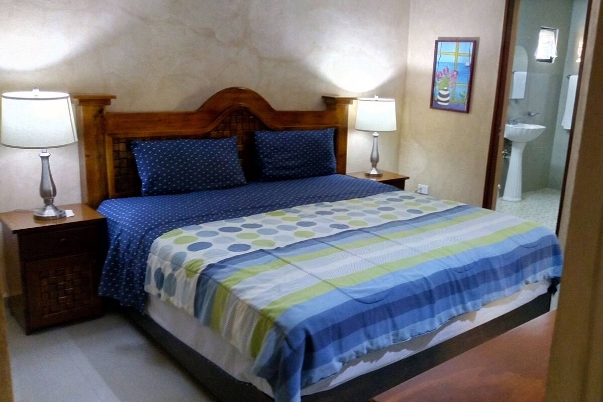Un dormitorio con cabecera de madera, dos lámparas de noche, una cama con colcha a rayas azules y verdes, y un cuadro en la pared.