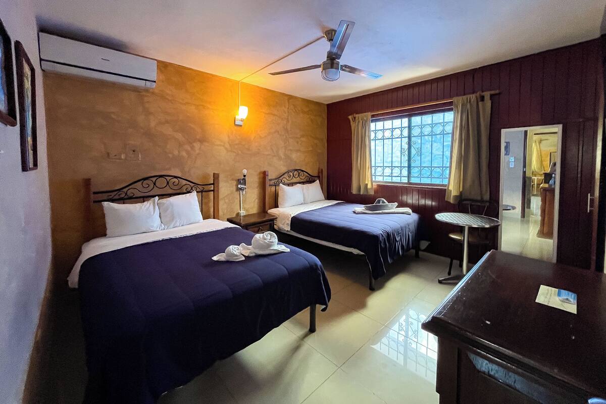 Habitación de hotel con dos camas, ventilador de techo, ventana con cortinas, una mesita y un espejo.