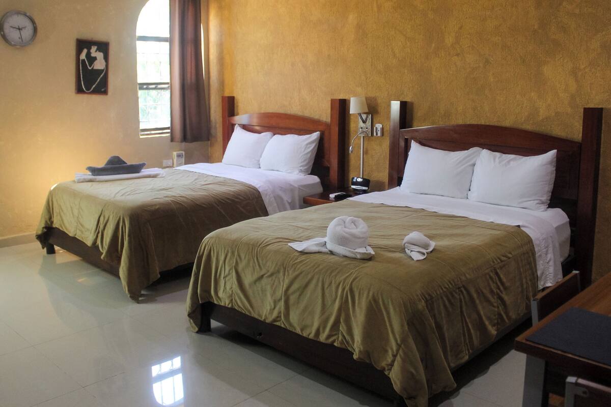Habitación de hotel con dos camas, un reloj, una ventana con cortinas y una mesita de noche con lámpara.