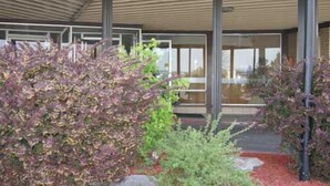 A modern building with a covered entrance, large windows, and a landscaped garden with various shrubs.