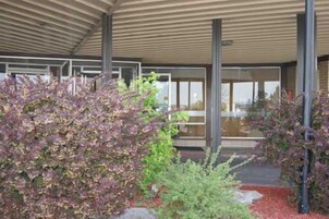Un edificio moderno con un acceso cubierto, amplios ventanales y un jardín ajardinado con diversos arbustos.