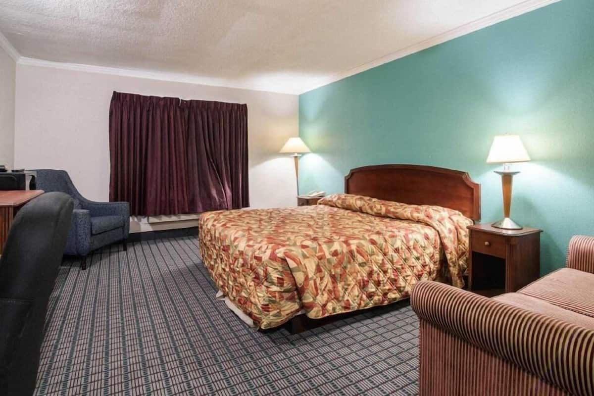 Quarto de hotel com cama, duas luminárias de cabeceira, escrivaninha com cadeira, janela com cortinas e tapete estampado.