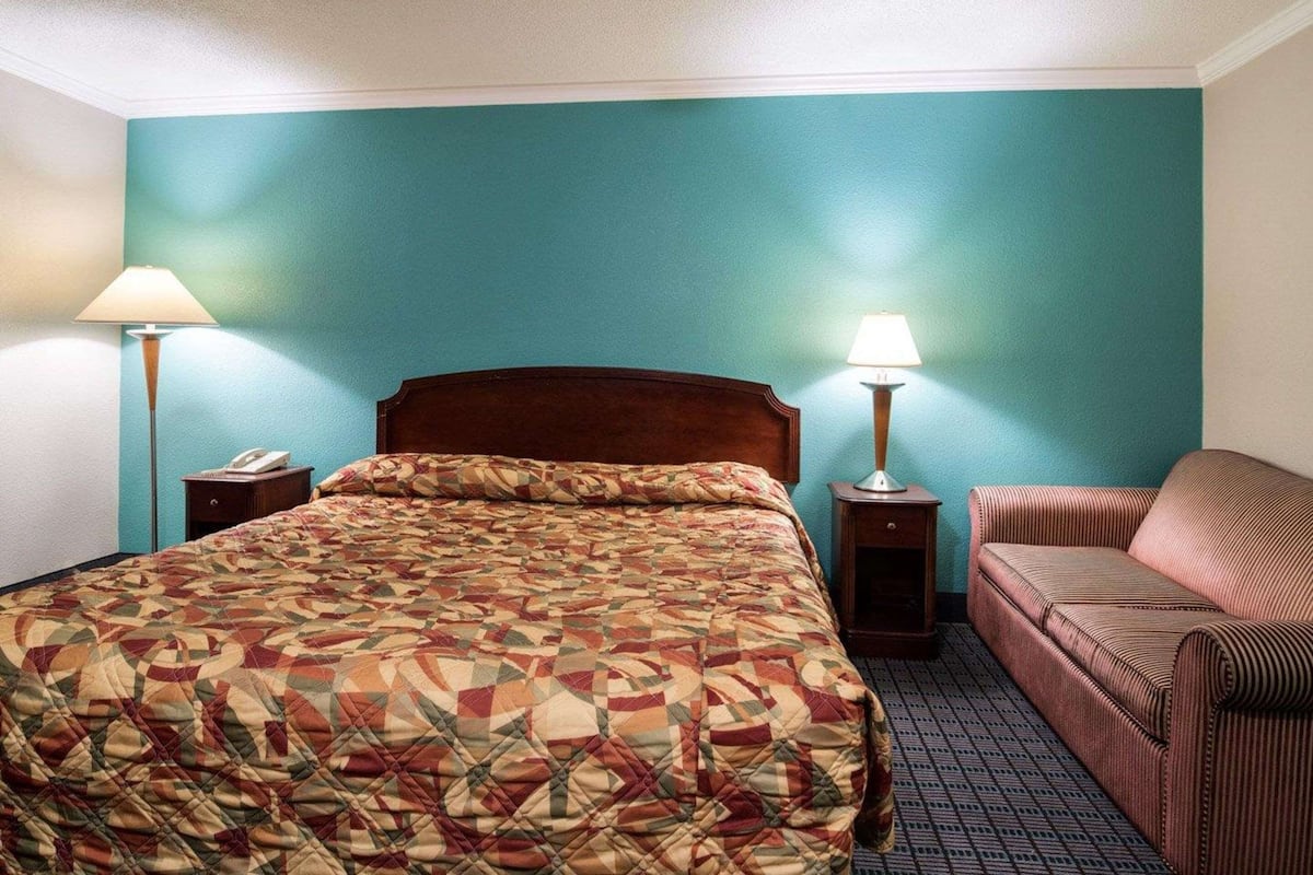 Quarto de hotel com cama, duas luminárias de cabeceira, telefone, sofá e tapete estampado.
