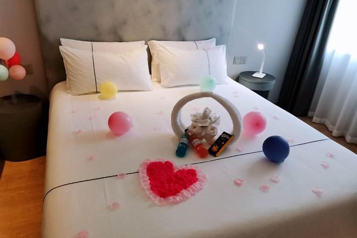 A neatly made bed with white bedding, a heart-shaped decoration, and a small table with a bottle and a candle.