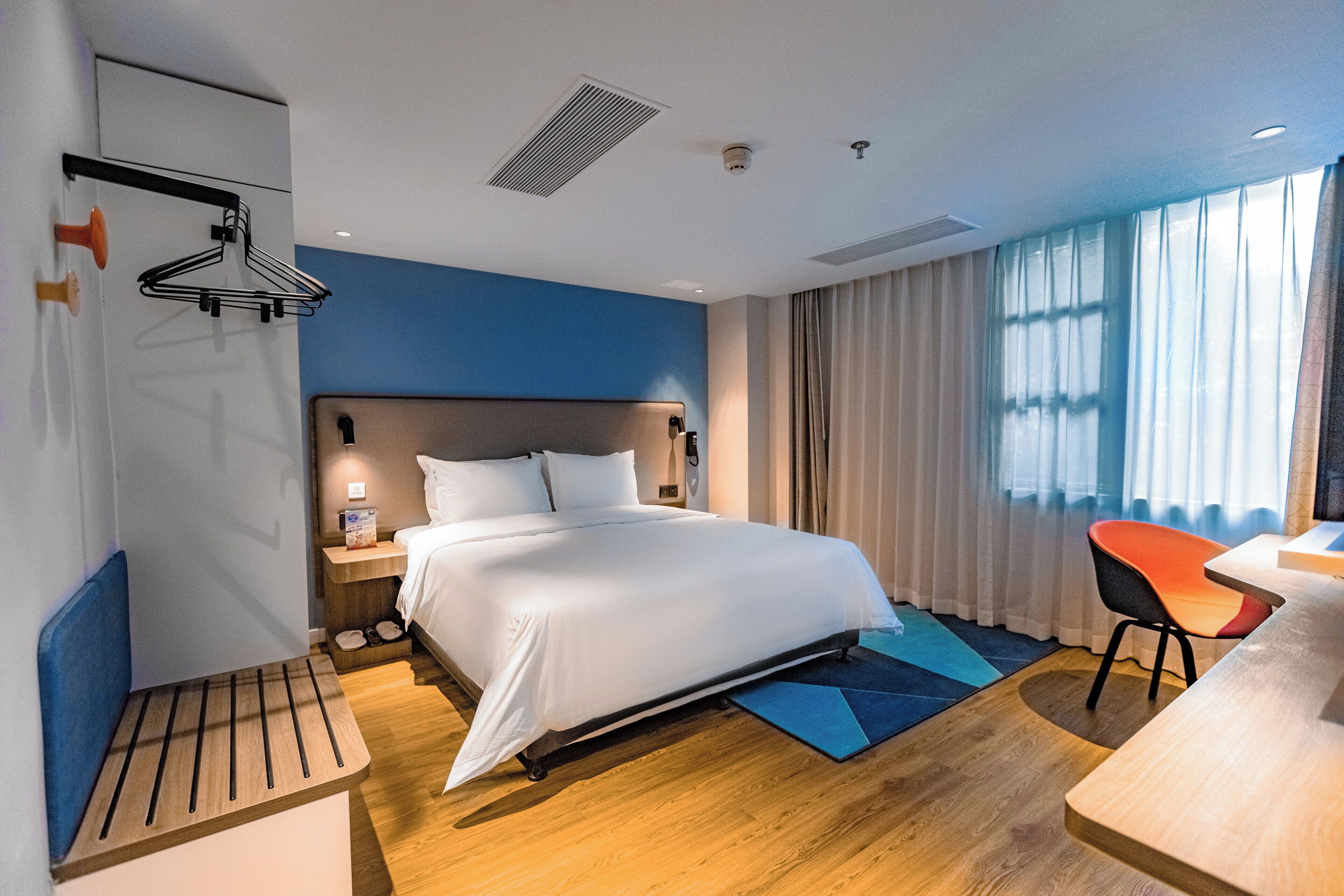 A modern hotel room with a large bed, bedside table, and a desk with a chair.