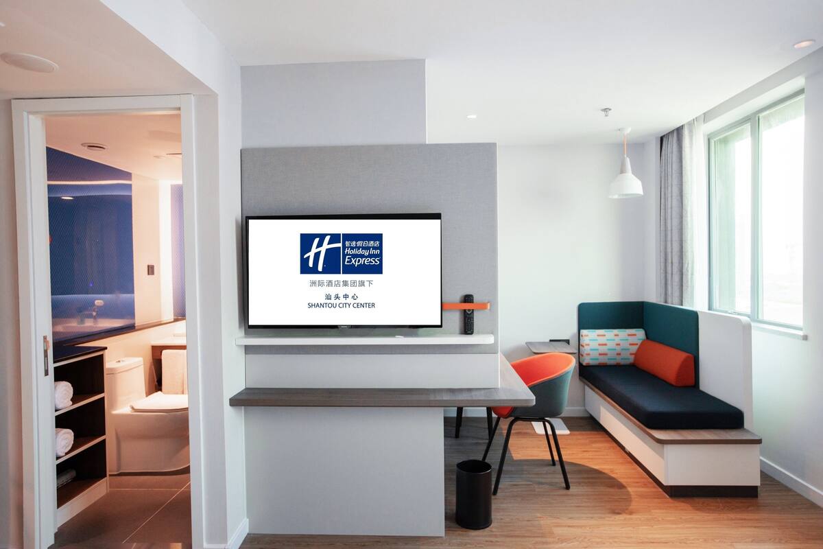Ein modernes Hotelzimmer mit einem großen Fernseher, der das Holiday Inn Express Logo zeigt, einem Sitzbereich mit Sofa und Sessel sowie Blick auf die Innenstadt durch das Fenster.