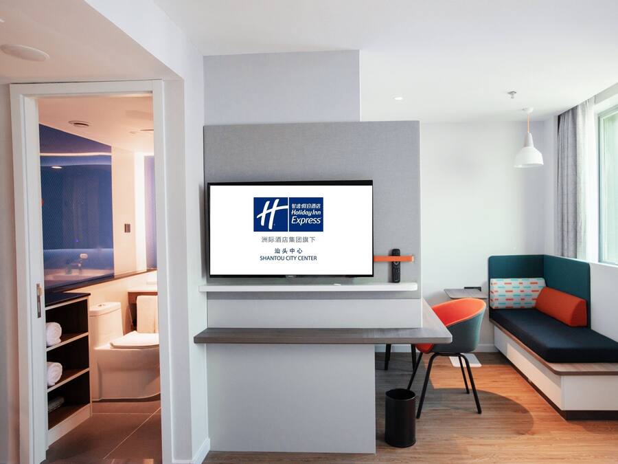 A modern hotel room with a large TV screen displaying the Holiday Inn Express logo, a seating area with a sofa and a chair, and a view of the city center through the window.
