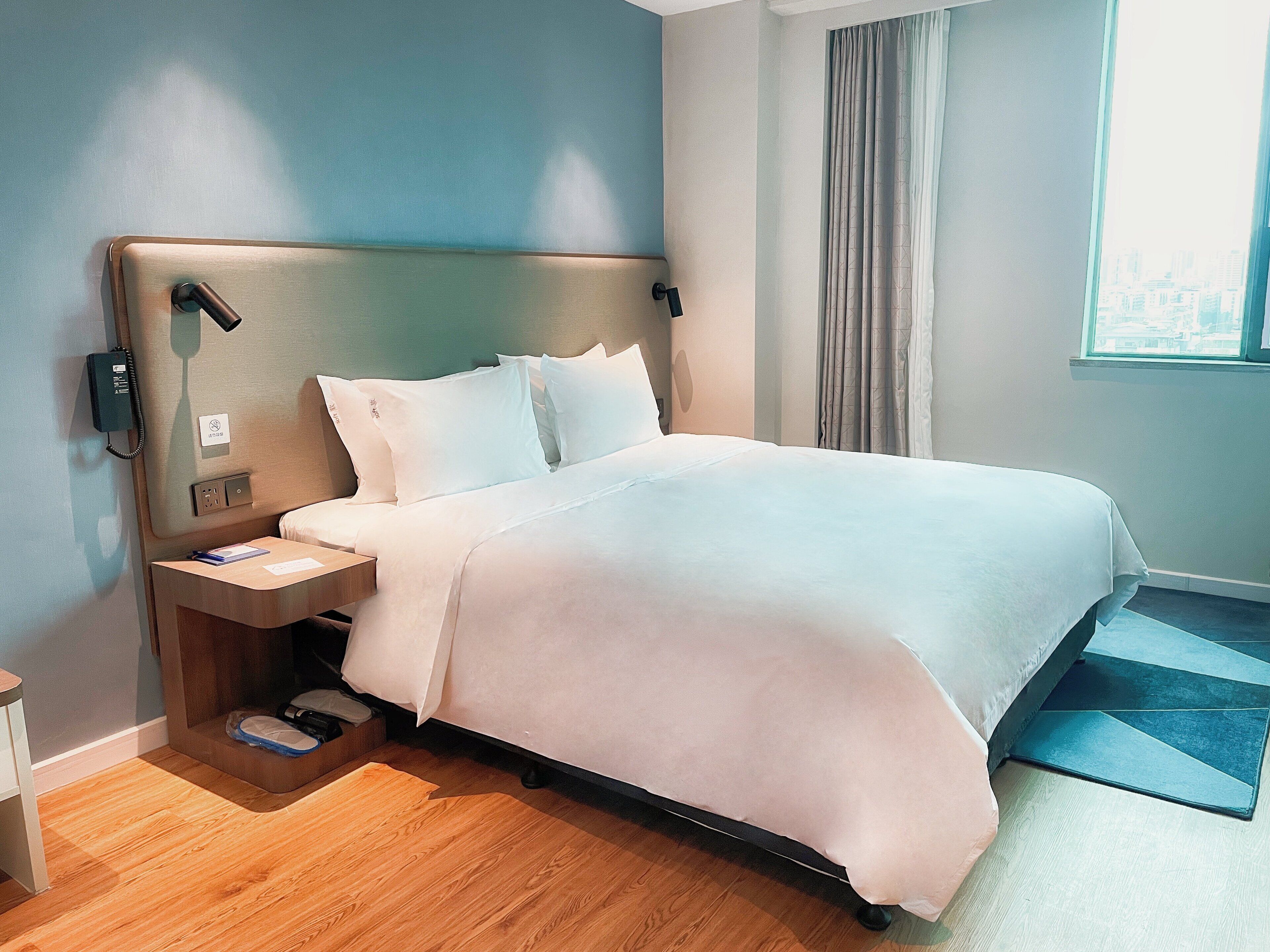 A hotel room with a bed, bedside table, and a window with curtains.