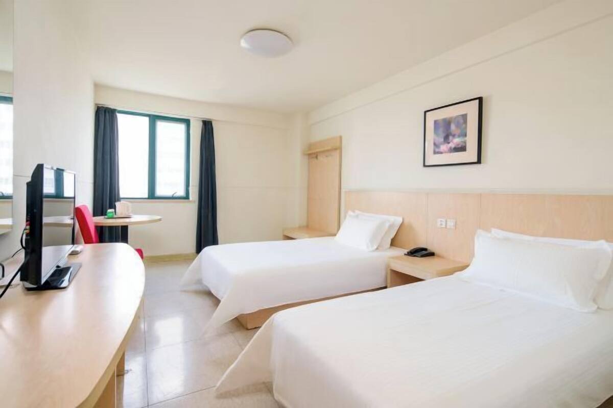 Hotelkamer met twee bedden, een bureau, een stoel, een klein tafeltje, een televisie en een raam met gordijnen.
