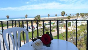 A balcony with a white chair and a table with two roses, overlooking a beach with palm trees and a road with cars. - Hotel Oasis (Conil de la Frontera)