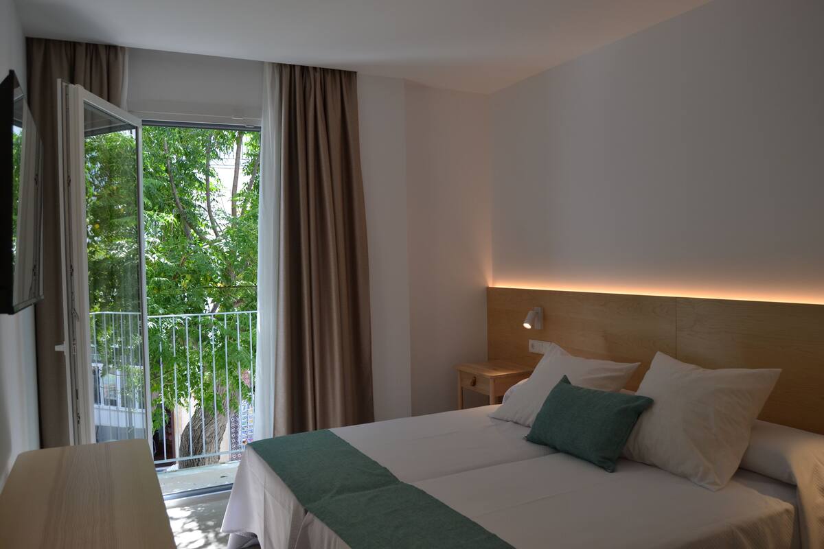 A hotel room with a bed, a nightstand, a lamp, and a view of greenery through a window.