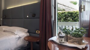 A hotel room with a bed, a bedside table with a tray of toiletries, and a window with a view of greenery outside. - Manuelina Taste Hotel (Recco)
