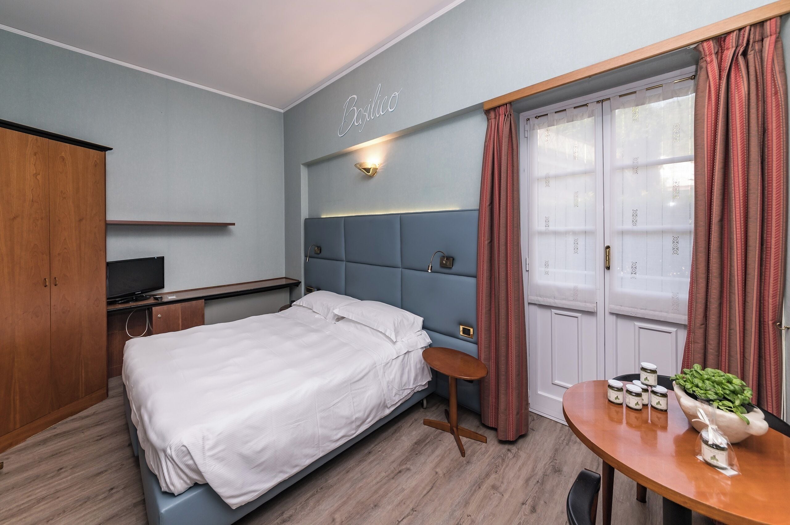 A bedroom with a bed, a desk, a chair, a TV, a wardrobe, and a window with curtains.