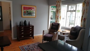 Ein Wohnzimmer mit einem dunklen Holztisch, einem gemusterten Teppich, einem Sessel, einem Sofa, einer Kommode und einem Fenster mit Vorhängen.