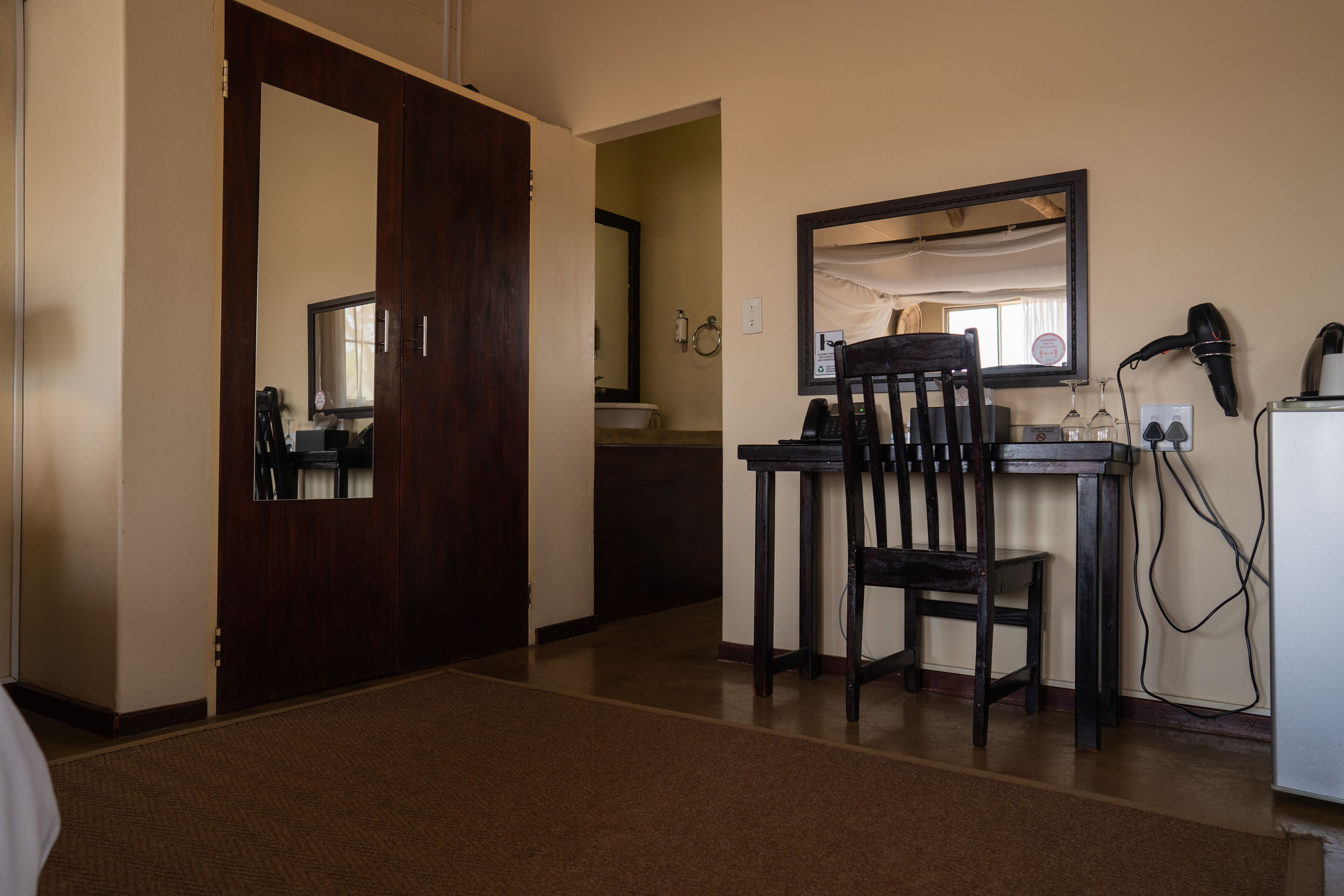 Une chambre d’hôtel avec une porte en bois, un bureau avec une chaise, un miroir, un sèche-cheveux et un réfrigérateur.