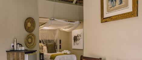 A room with a bed, a desk, and a chair. There is a picture on the wall and a mosquito net over the bed.