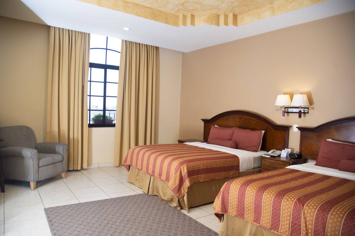 Ein Hotelzimmer mit zwei Betten, einem Sessel, einem Nachttisch und einem Fenster mit Vorhängen.