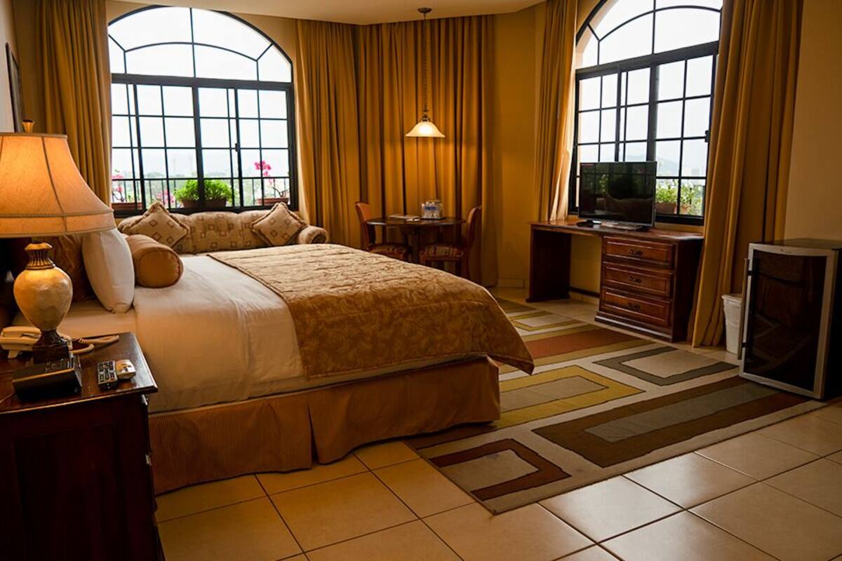 Ein Hotelzimmer mit einem großen Bett, einem Schreibtisch mit Lampe, einem Fernseher und Blick durch große Fenster.