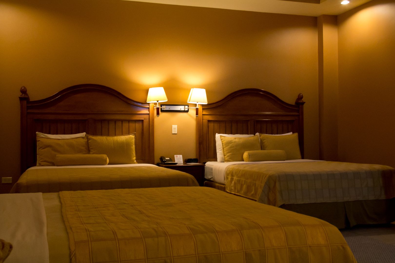 A hotel room with two beds, each with a wooden headboard and matching pillows. There are two bedside lamps and a nightstand with a phone and a notepad.