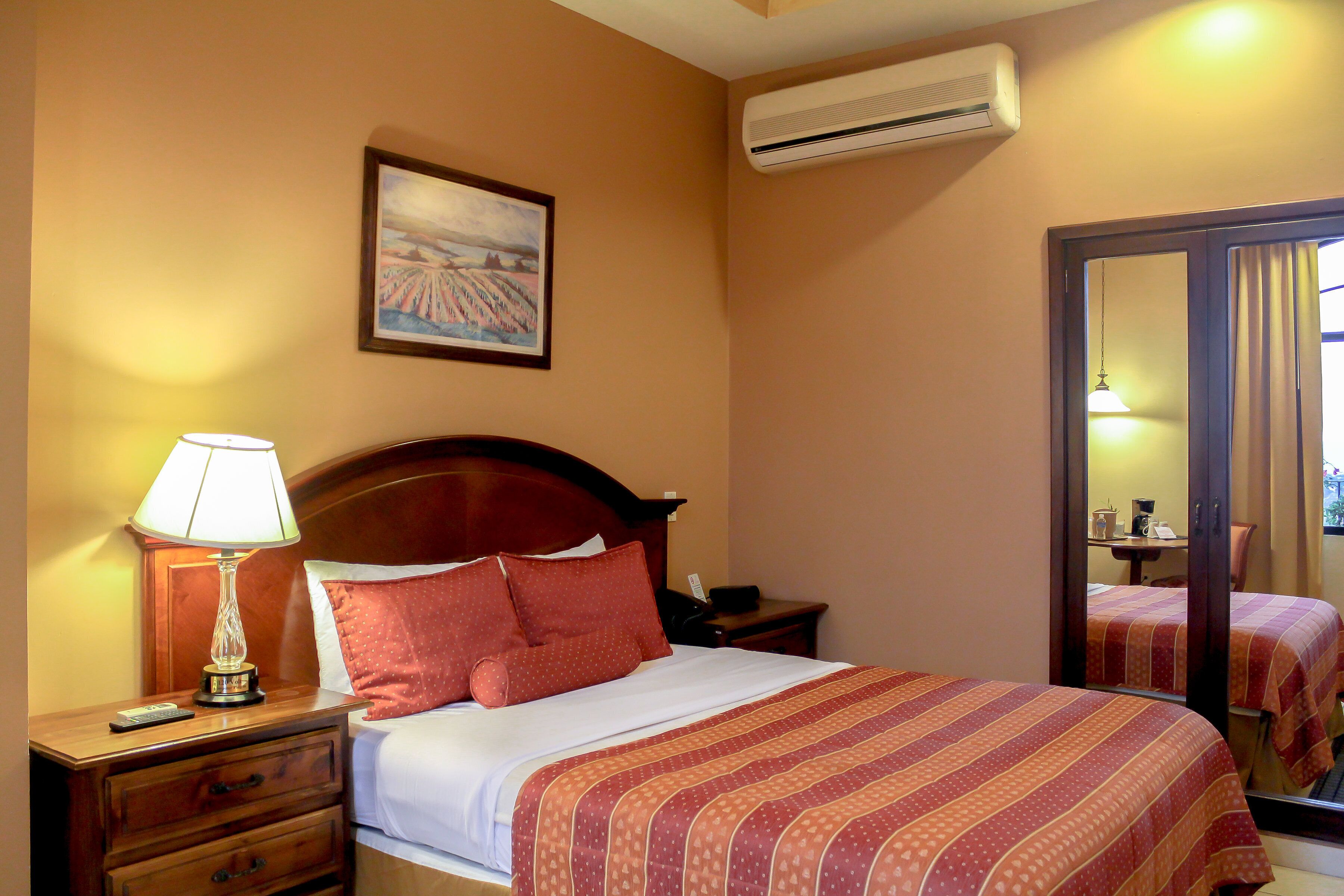 Habitación de hotel con cama, mesita de noche con lámpara, un cuadro en la pared y un equipo de aire acondicionado.