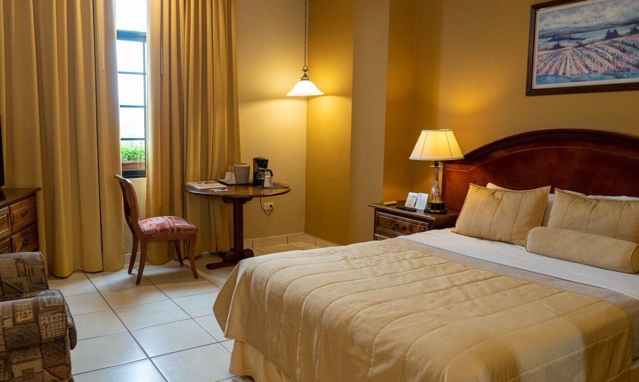 Una habitación de hotel con una cama grande, un escritorio con lámpara, una silla y un ventanal con cortinas.