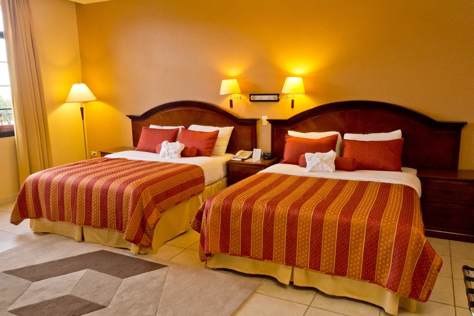 A hotel room with two beds, each with a striped bedspread, a nightstand with a lamp, and a wall-mounted telephone.