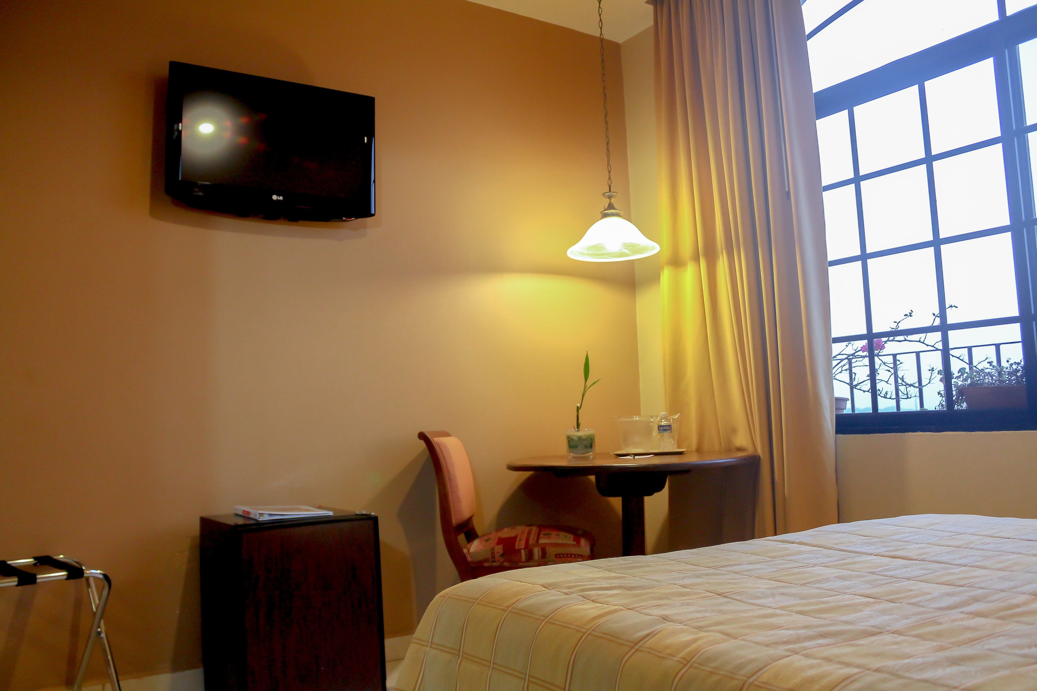 Habitación de hotel con cama, televisor montado en la pared, mesa con silla y ventana con cortinas.