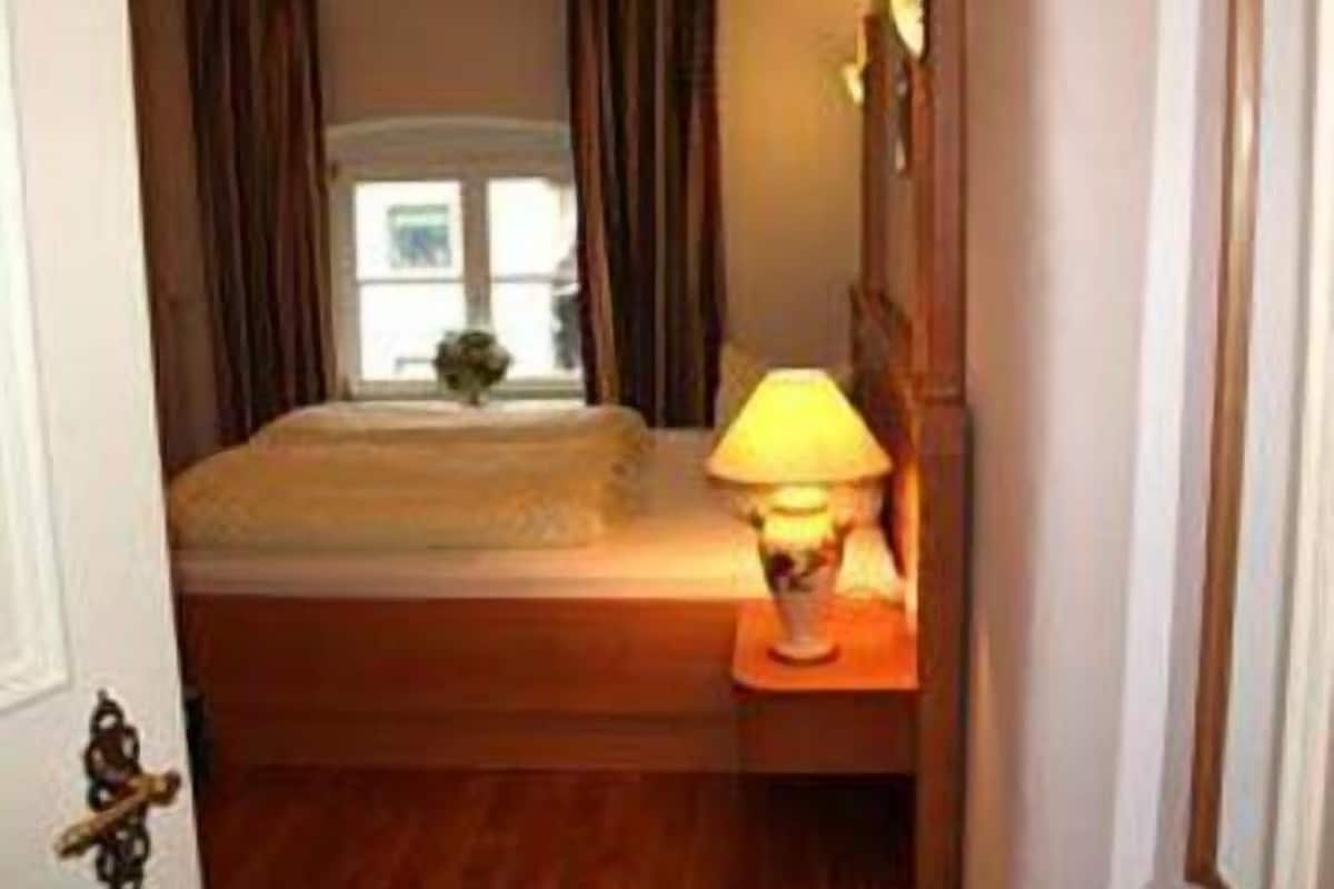 Ett litet hotellrum med en säng, ett nattduksbord med en lampa och ett fönster med gardiner.