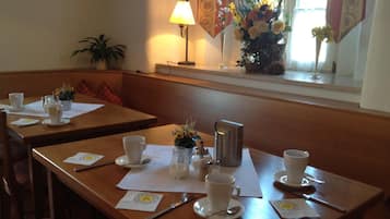 A dining area with tables set for two, each with a cup, saucer, spoon, and napkin. There is a lamp, a vase with flowers, and a potted plant on the windowsill.