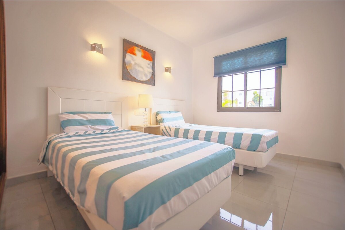 A hotel room with two beds, a window with blinds, a wall-mounted light, and a painting.