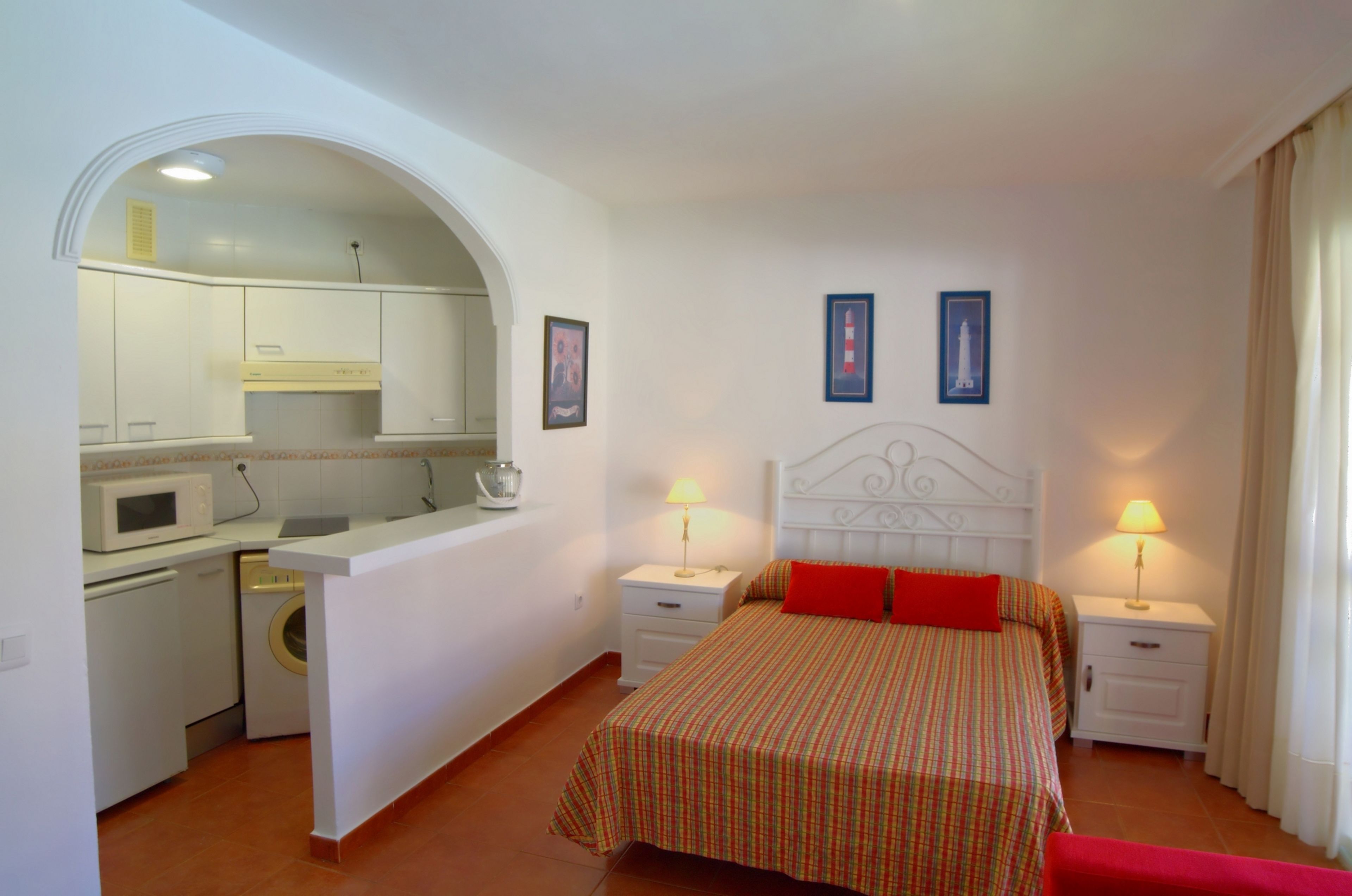 Habitación de hotel compacta con kitchenette, una cama con almohada decorativa roja, dos lámparas de noche y un baño pequeño.