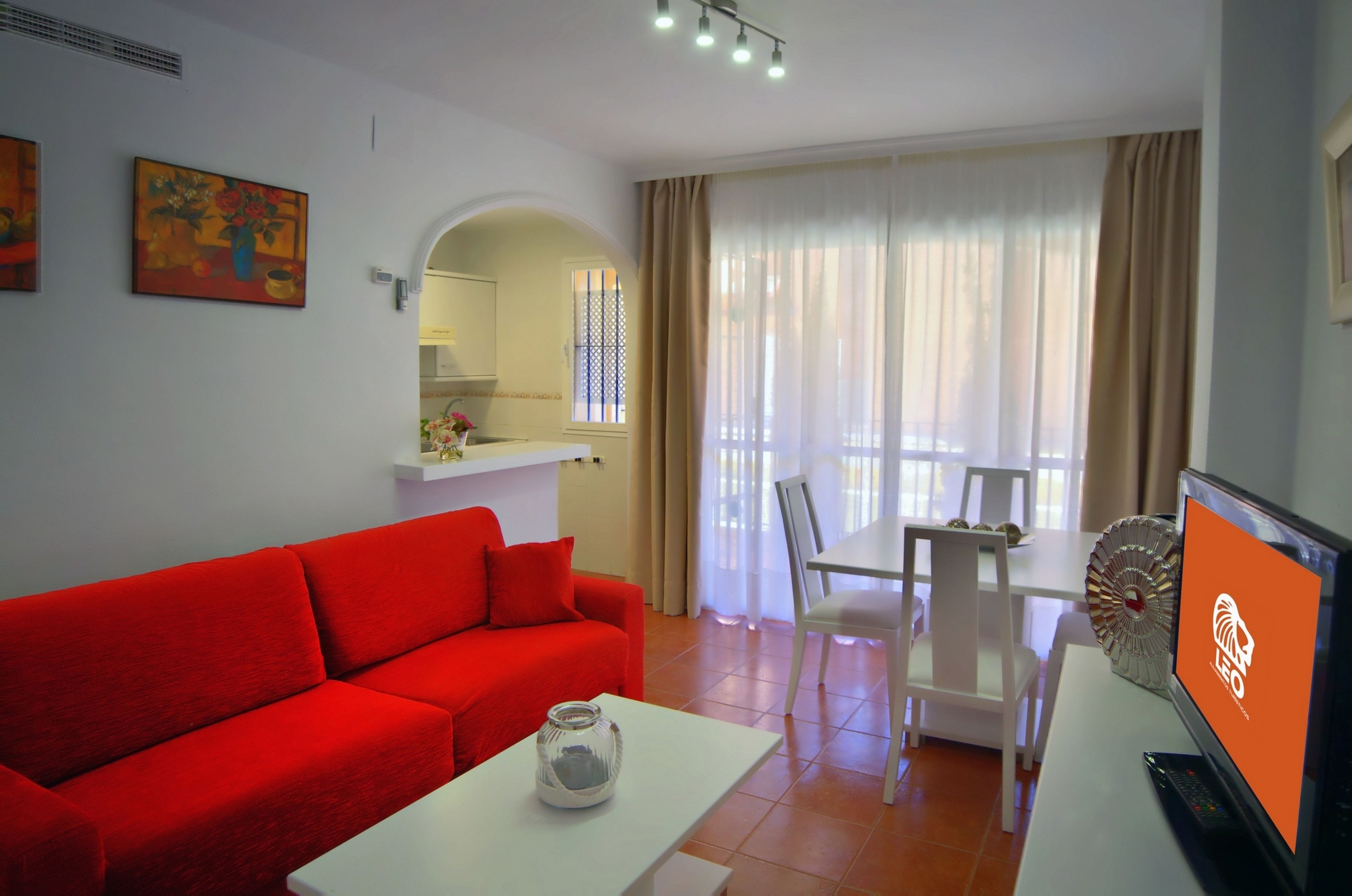 Una sala de estar moderna con un sofá rojo, una mesa de centro blanca y un comedor con una mesa y sillas blancas. Hay un televisor sobre un mueble, con un logotipo en la pantalla.