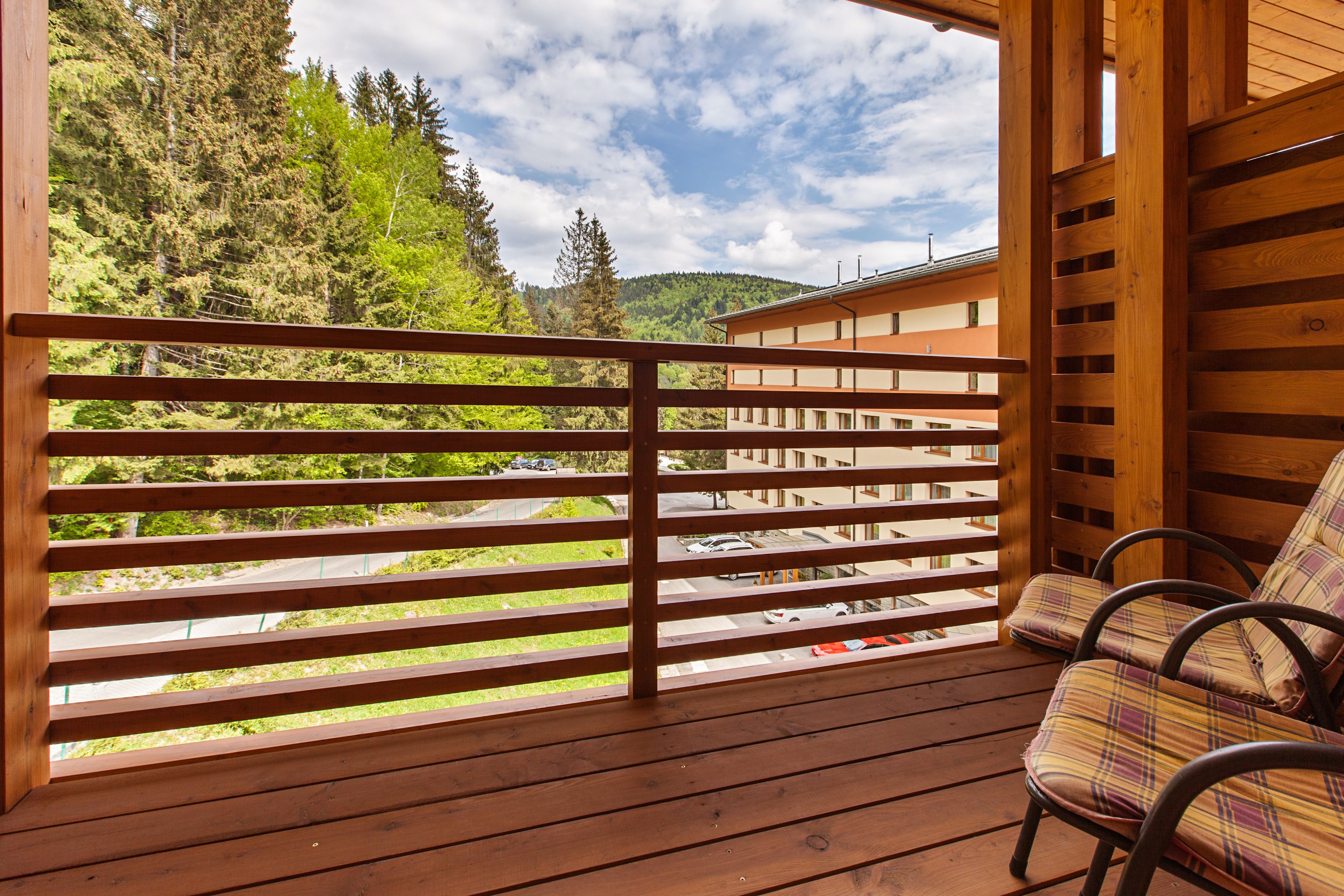 A wooden balcony with a view of a forest and a building. There are two chairs with plaid cushions.