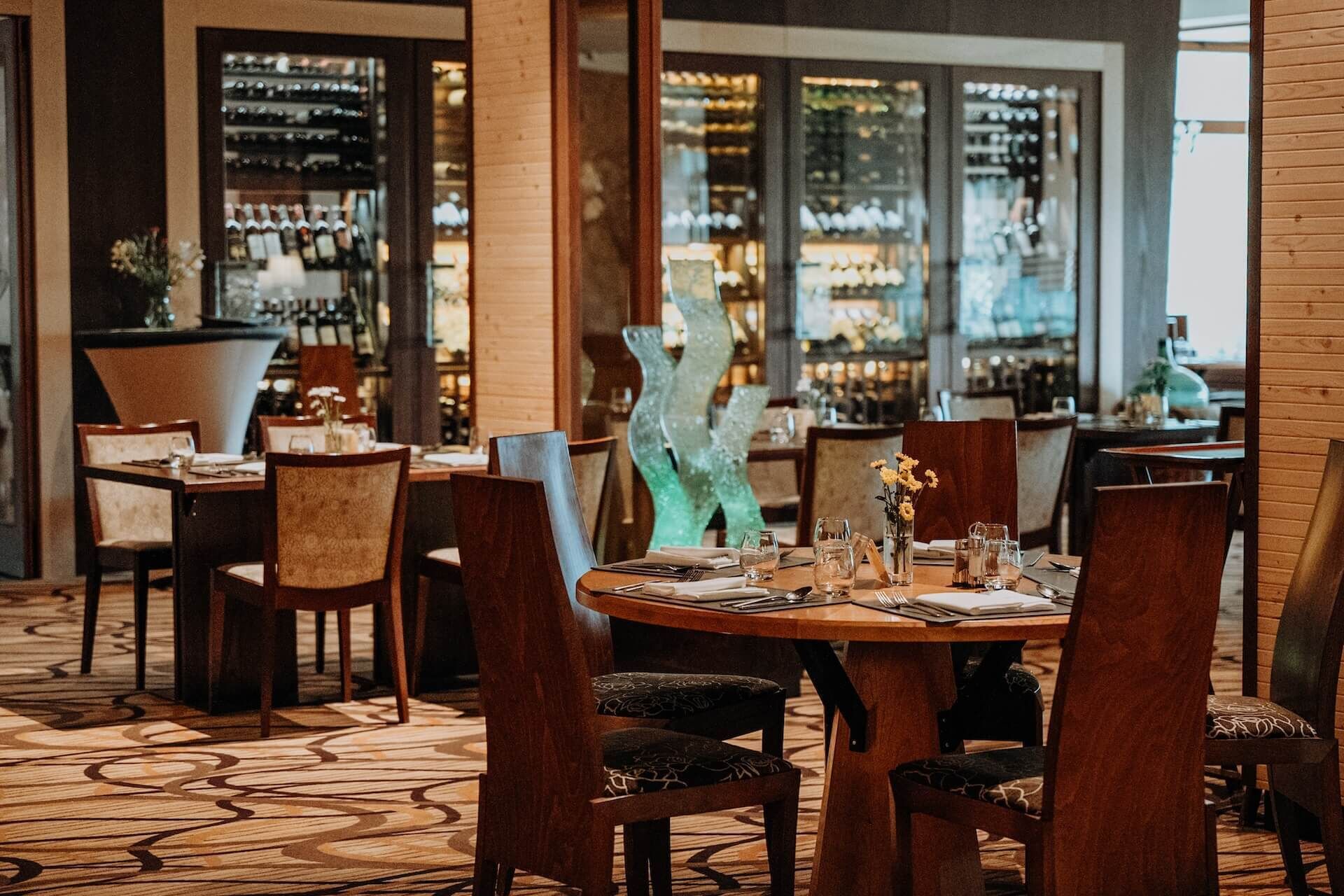 En restauranginteriör med runda bord, trästolar och en bardisk som syns genom glasväggen.