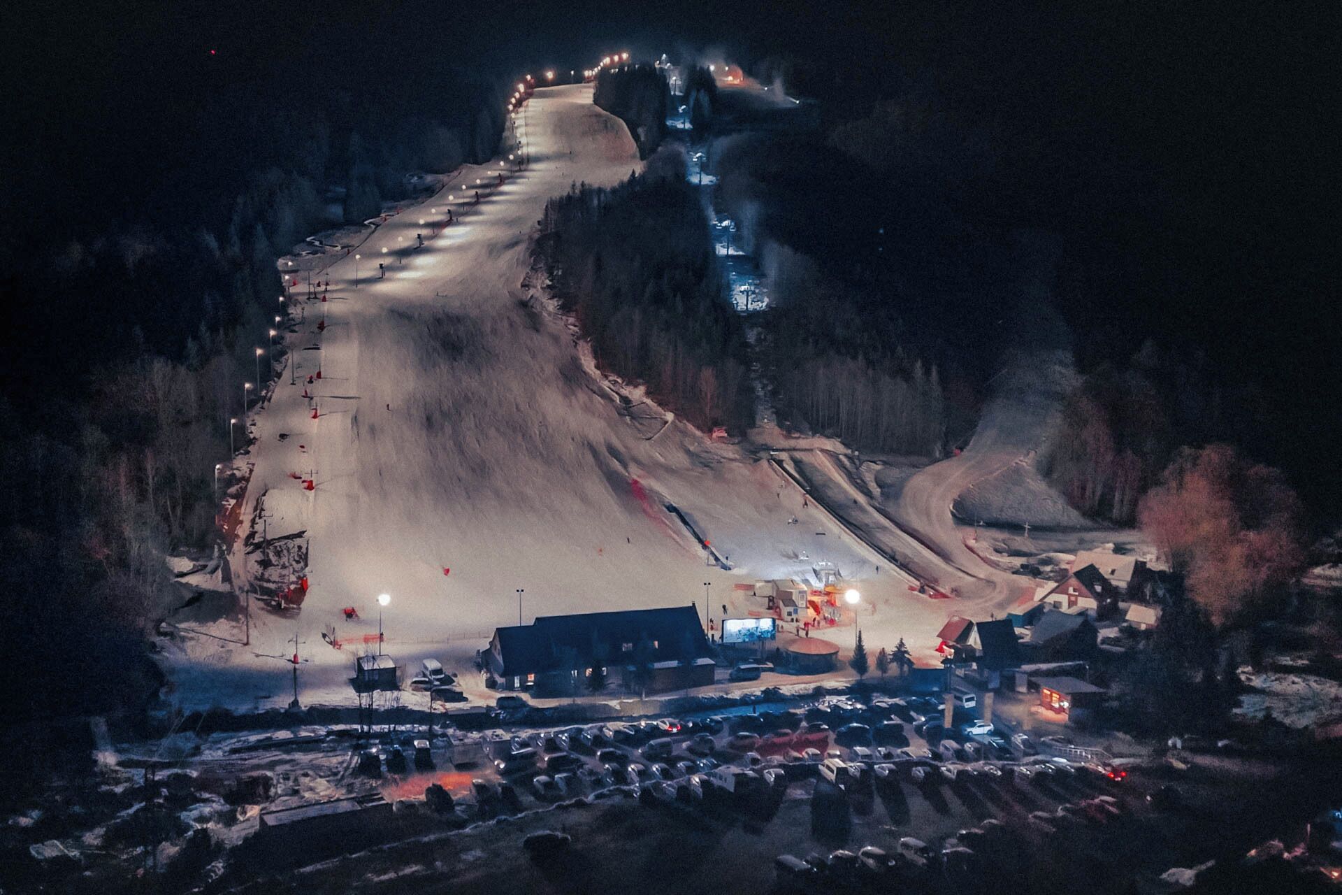 A snow-covered ski slope at night with illuminated ski lifts and a parking area at the base.