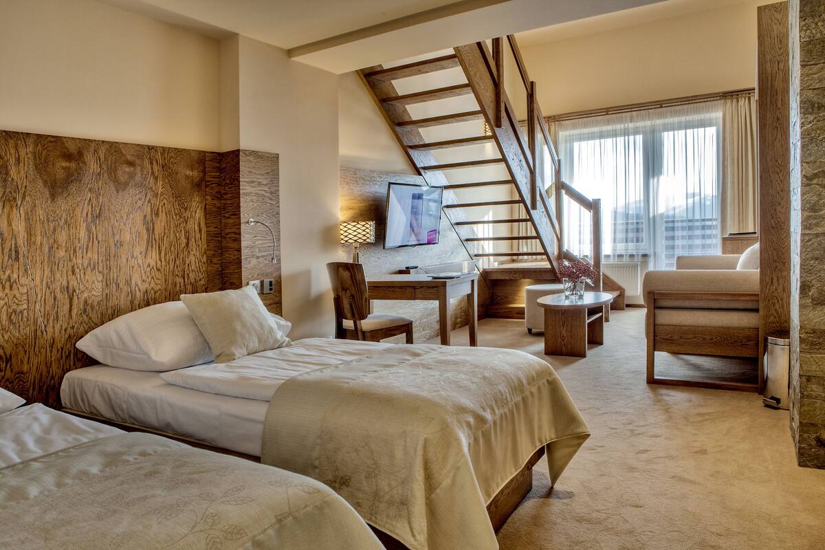 A hotel room with two beds, a desk, a chair, a small table, a TV, and a staircase.