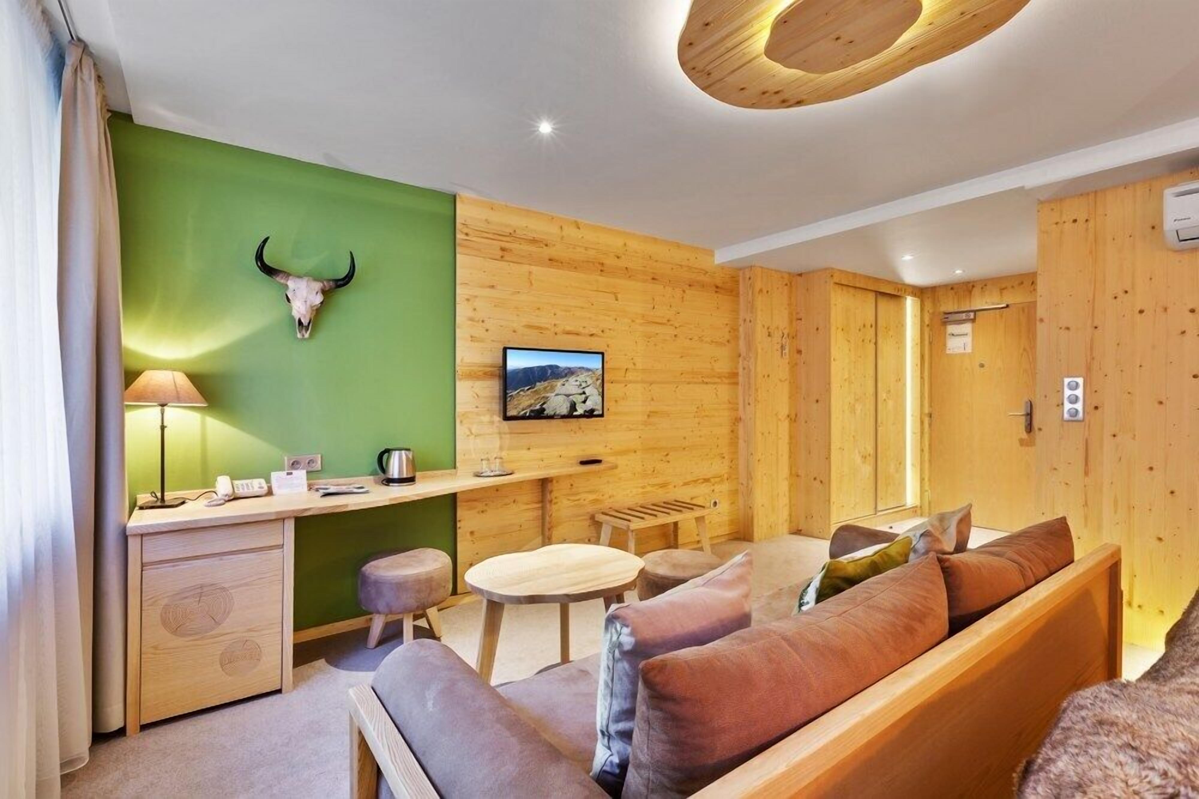 Ein gemütliches Zimmer mit einem Holztisch, einer grünen Akzentwand, einem aufgehängten Kuhschädel, einer Couch und einer Holzzellentrale.