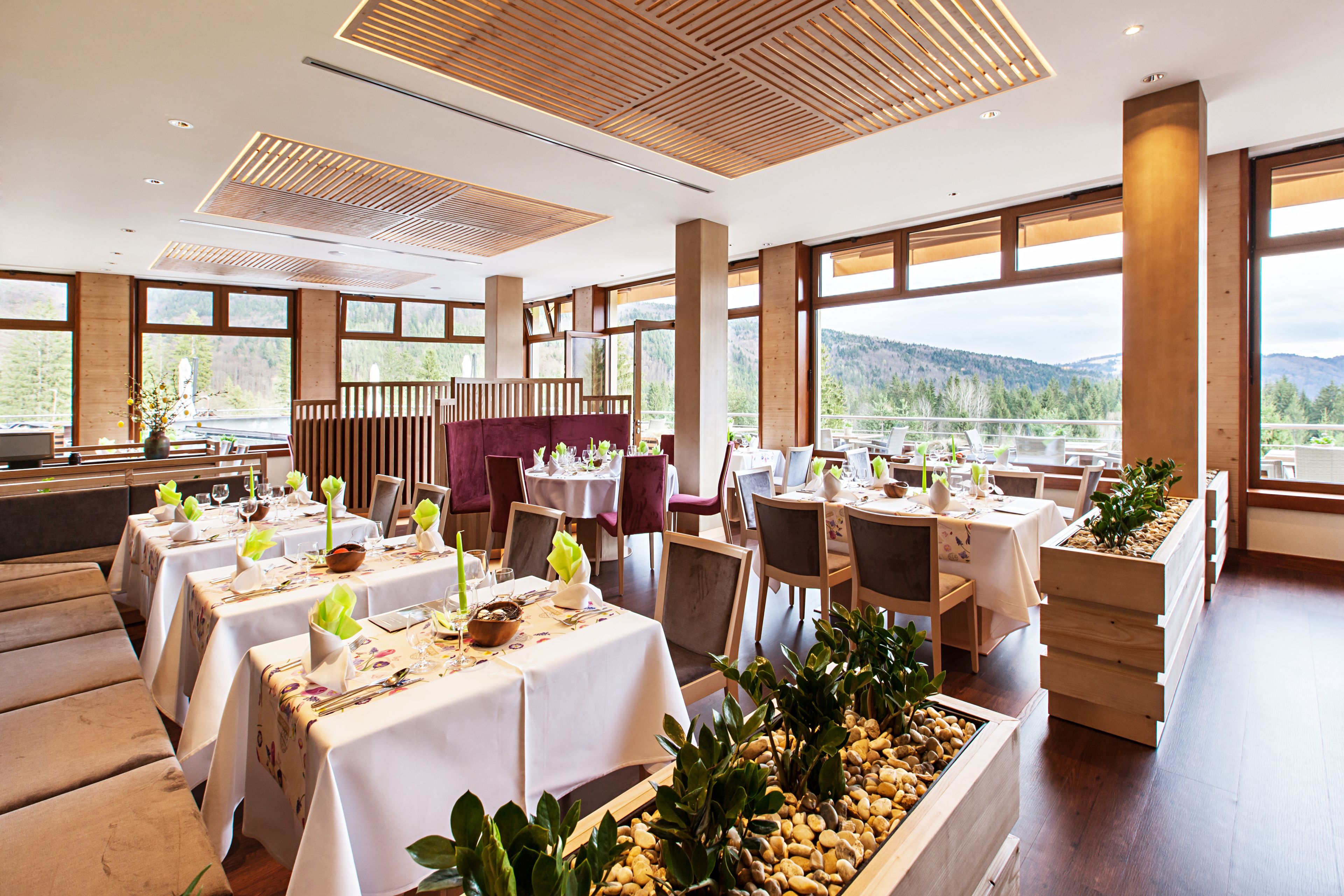 A restaurant with multiple tables set for dining, large windows offering a view of the outdoors, and a modern ceiling design.