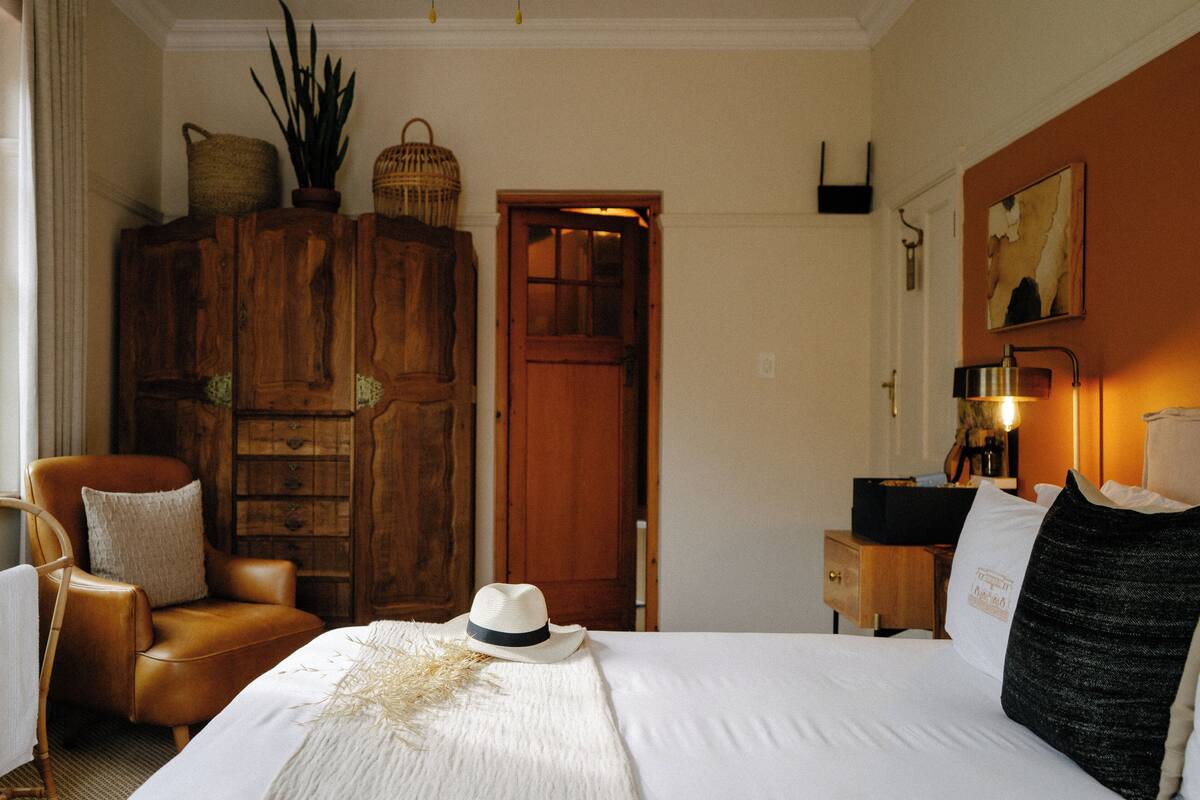 A bedroom with a wooden wardrobe, a bed with white bedding, a leather armchair, a bedside table with a lamp, and a painting on the wall.