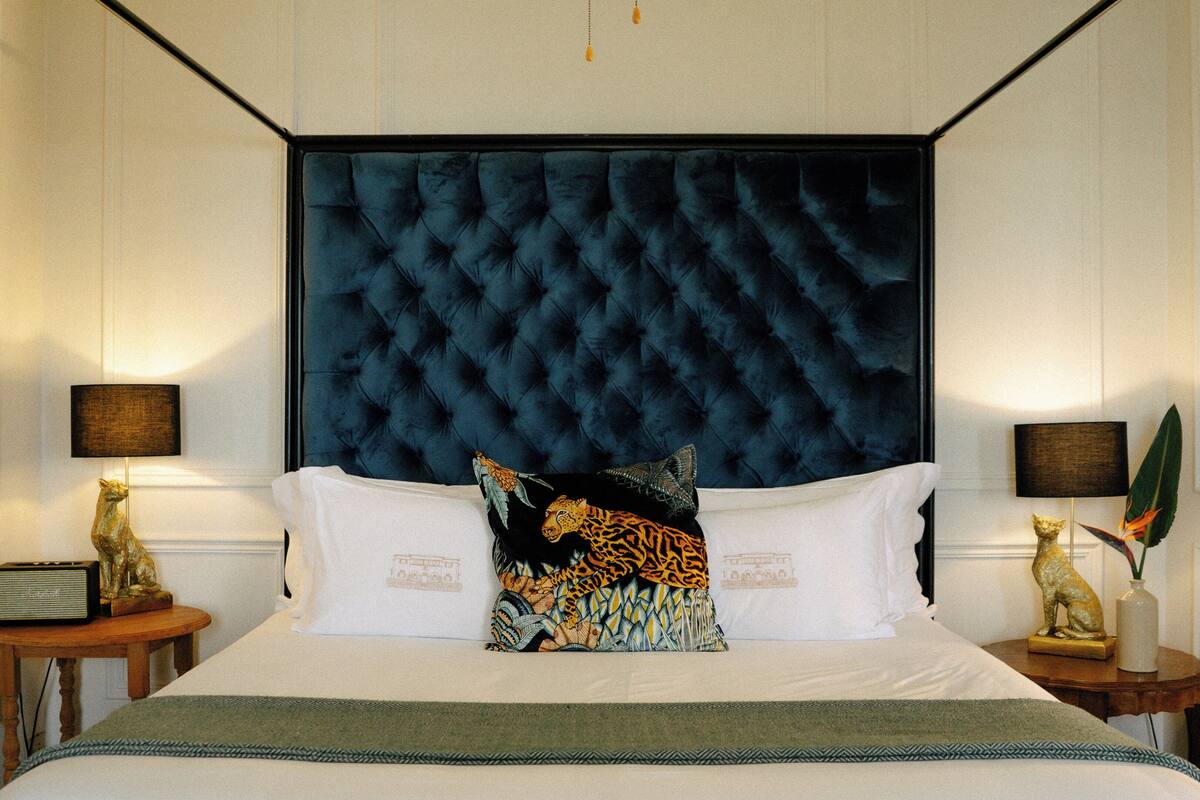 A hotel room with a tufted headboard, a bed with white linens, two nightstands with lamps and decorative items, and a green throw blanket.