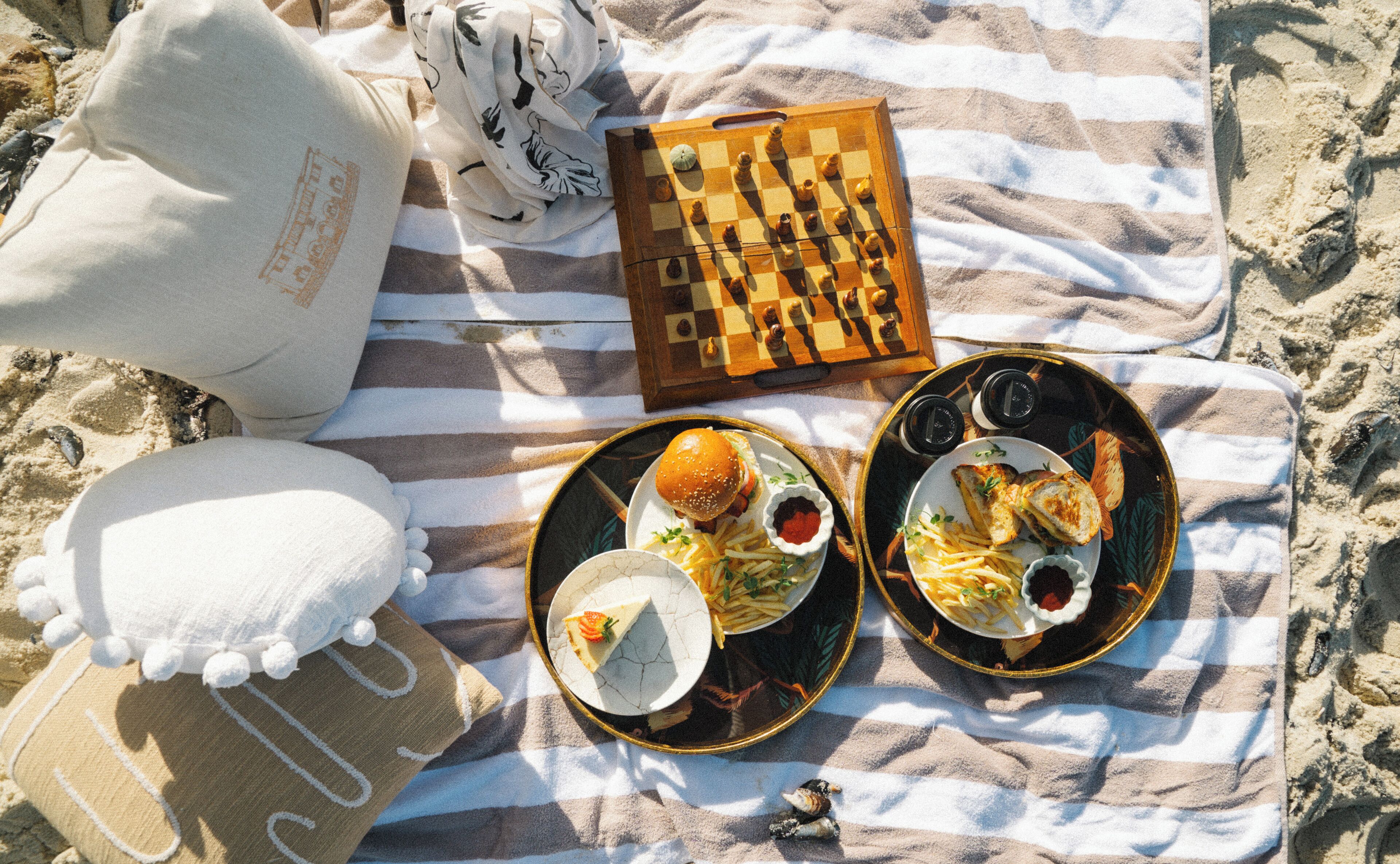 A beach scene with a chessboard, two plates of food, and a white pillow on a striped beach towel.