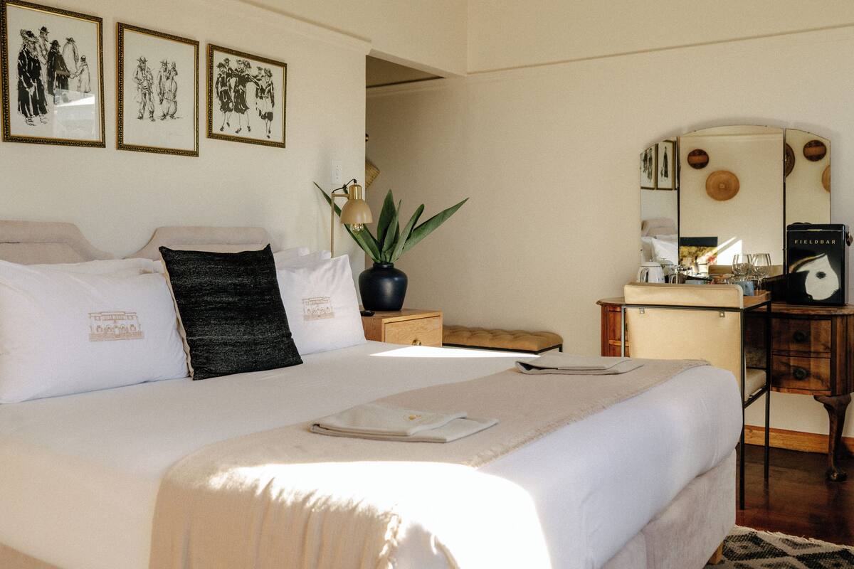 A neatly made bed with white linens and a black pillow, a small wooden nightstand with a lamp, a mirror, and a Fieldbar coffee machine.