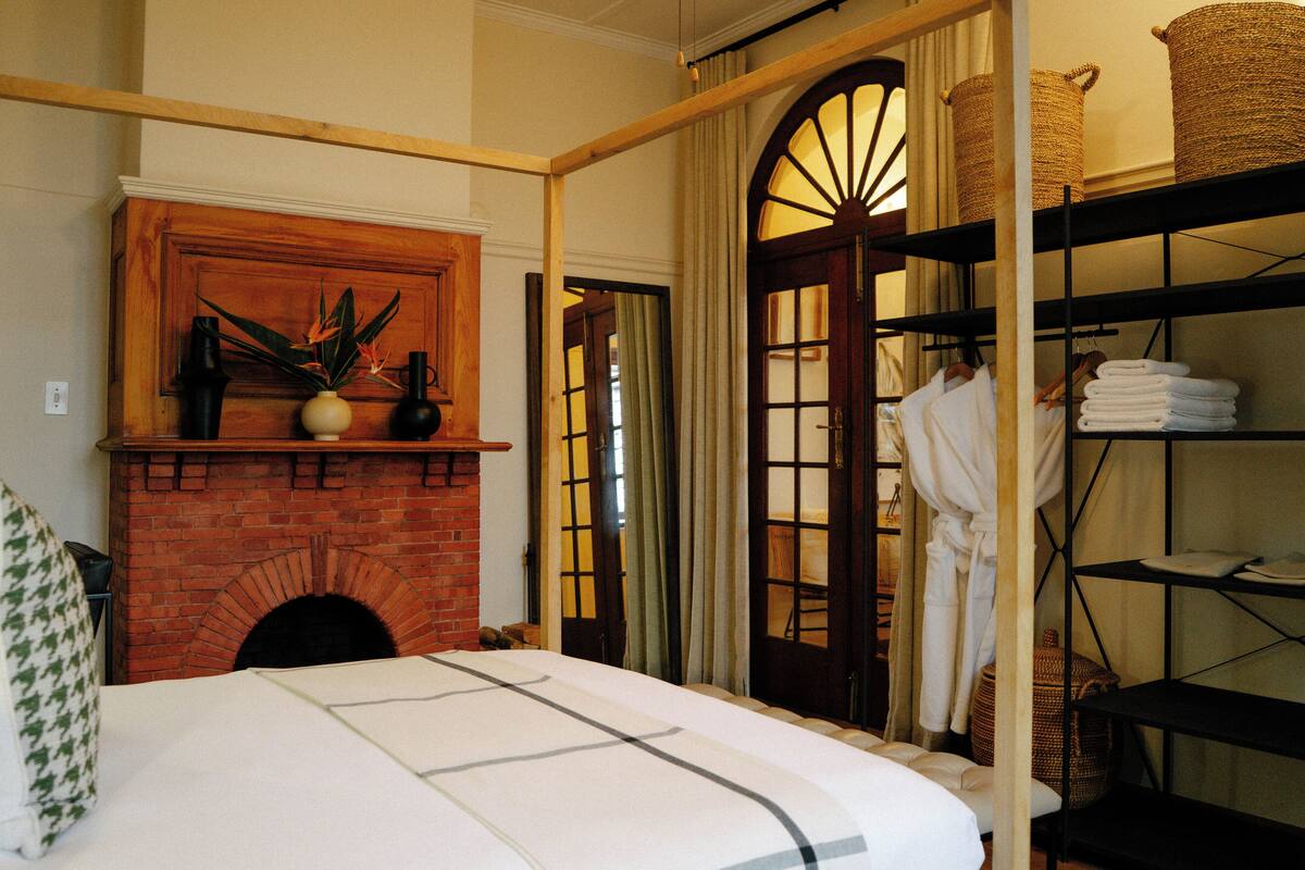 A hotel room with a brick fireplace, a bed with a white and black striped bedspread, a wooden shelf with towels, and a large arched doorway.