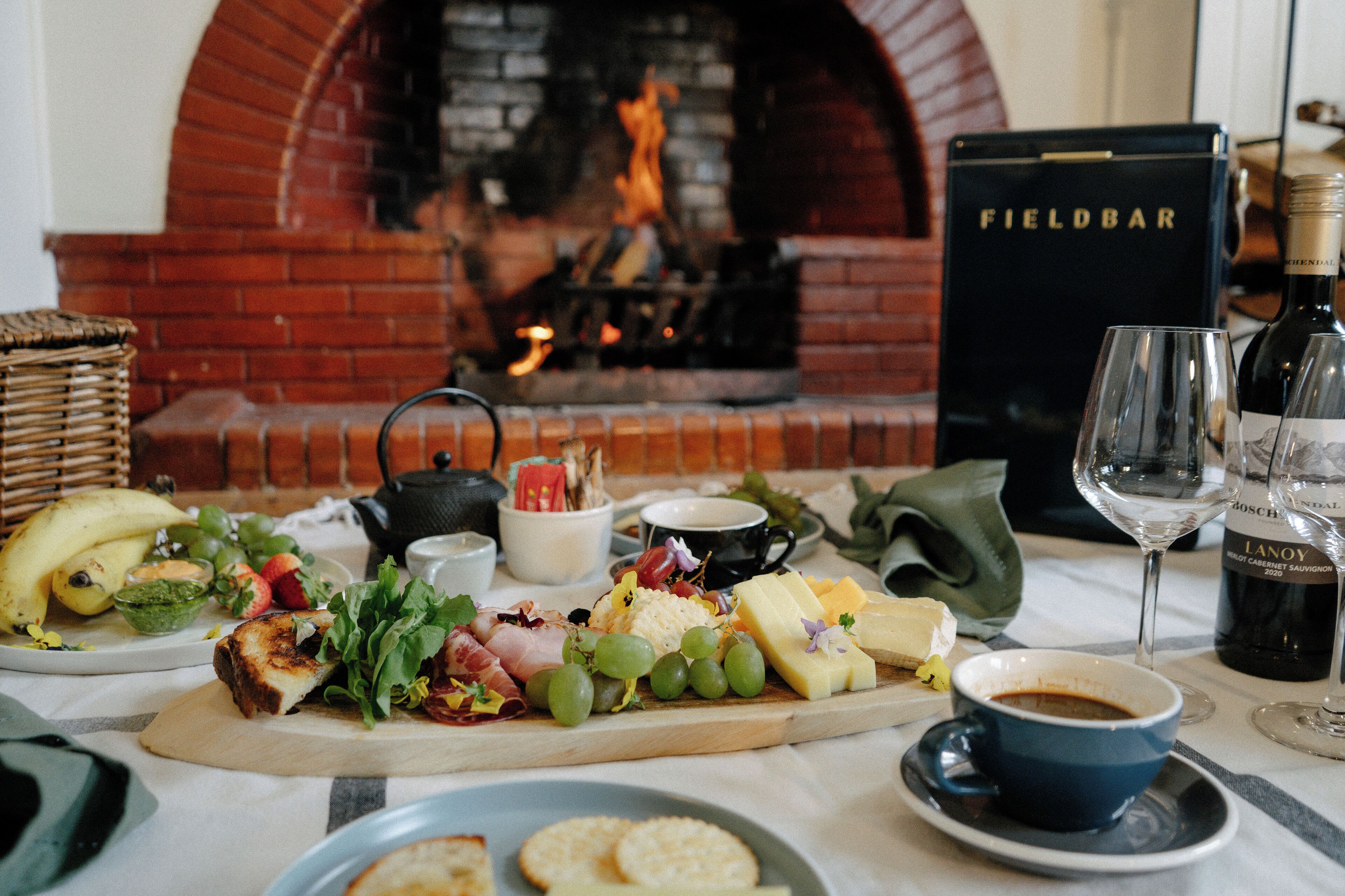A table set with a cheese board, fruits, and beverages, with a fireplace and a Fieldbar sign in the background.